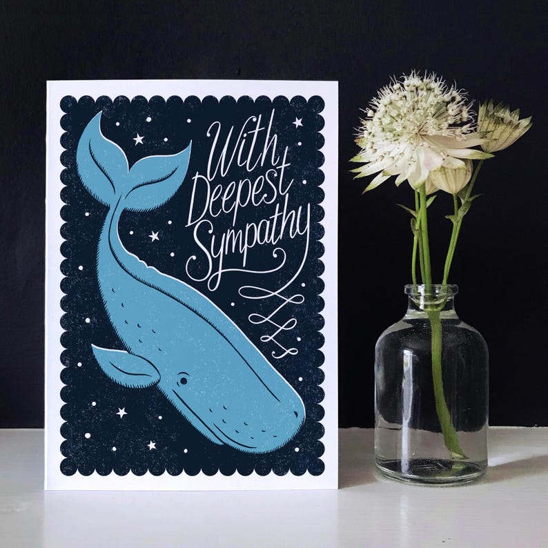 May include: A sympathy card featuring a blue whale illustration on a dark blue background with stars. The card reads "With Deepest Sympathy" in white script. A clear glass vase with white flowers is to the right.
