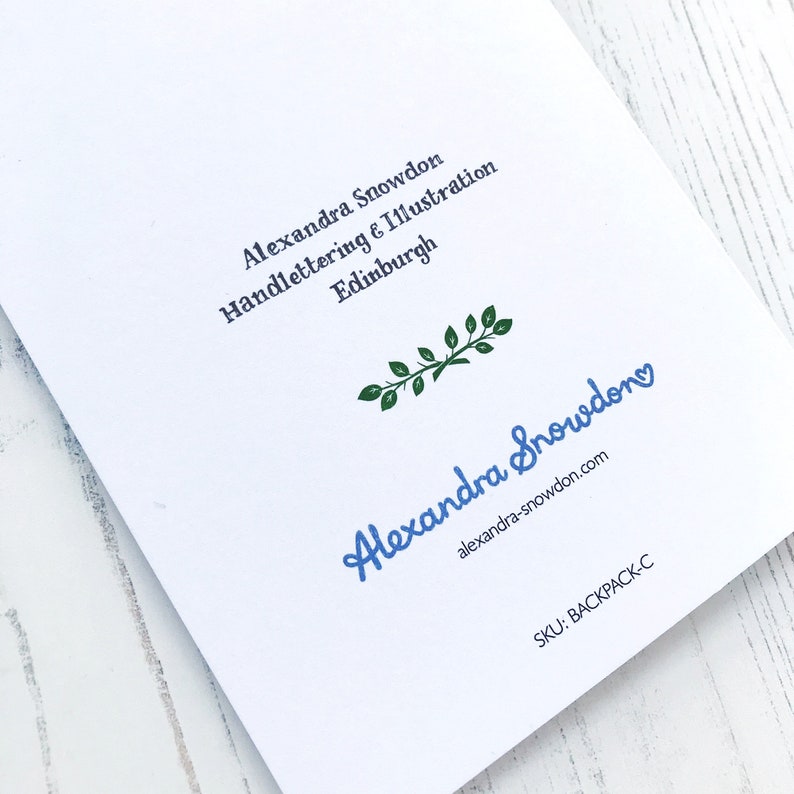 May include: A white card with blue text that reads "Alexandra Snowden Handlettering & Illustration Edinburgh" and a blue handwritten signature that reads "Alexandra Snowden" with a small heart above the "n".  The text "alexandra-snowdon.com" is printed in black below the signature.  A green leafy branch is above the signature.  The text "SKU: BACKPACK-C" is printed in black at the bottom of the card.