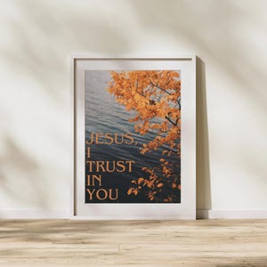 May include: Framed art print featuring the text "JESUS, I TRUST IN YOU" in a warm, golden hue. The image shows a body of water with a tree branch with orange leaves. The print is in a white frame.