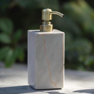 Könnte beinhalten: Ein rechteckiger Seifenspender aus weißem Marmor mit einer goldfarbenen Pumpe. Der Marmor hat eine dezente orangefarbene Maserung. Der Spender ist ein modernes Design, geeignet für Bad oder Küche.