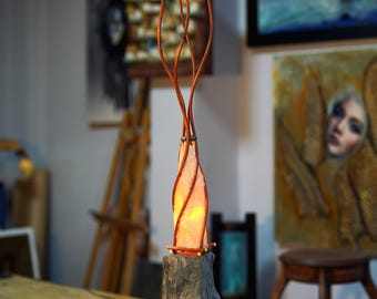 Puede incluir: Una lámpara de mesa con una base de madera y una pantalla de vidrio transparente retorcida. La pantalla está iluminada desde el interior, creando un brillo cálido. La lámpara está rodeada de ramas de madera, creando una estética natural y rústica.