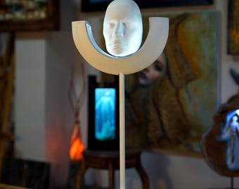 Puede incluir: Una escultura blanca de una cara que levita sobre una base blanca curva. La escultura está sobre un pedestal blanco con una base cuadrada.