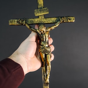 May include: A gold-toned crucifix with a figure of Jesus Christ on the cross. The crucifix is held in a person's hand.