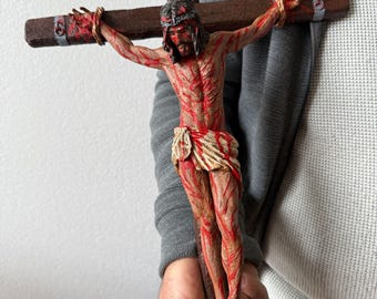 May include: A wooden crucifix with a detailed, realistic depiction of Jesus Christ. The figure is painted with red and white, representing blood and skin. The crucifix is held by a person's hand.