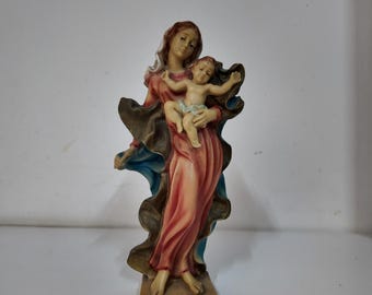 May include: A religious figurine depicting a woman in a flowing red and blue robe holding a child. The child is nude with arms outstretched. The woman has brown hair and a serene expression. The base is a rectangular shape.