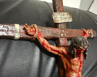 May include: A wooden crucifix with a depiction of Jesus Christ, showing a realistic portrayal of his wounds and blood. The crucifix is brown with a white inscription on the top.