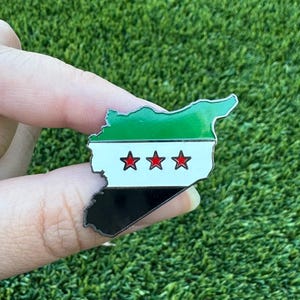 May include: A pin in the shape of the Syrian map, featuring the country's flag colors: green, white, black, and three red stars. The pin is held against a backdrop of green grass.