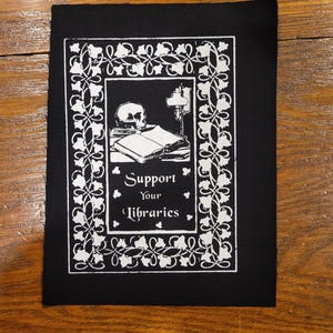 May include: Black patch with a white illustration. The design features a skull, books, and a lamp, framed by a decorative border. The text "Support Your Libraries" is centered below the illustration.