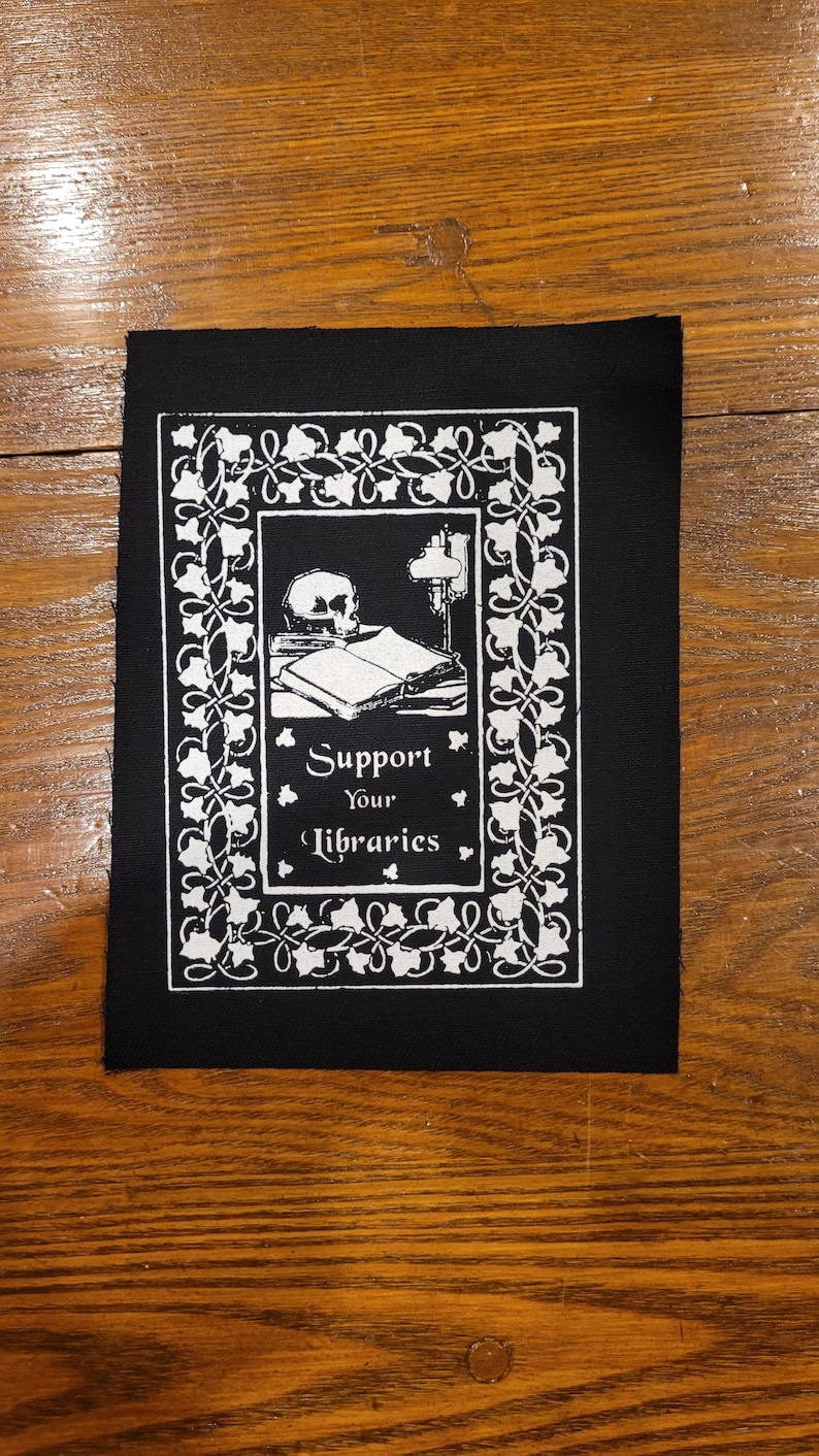 Puede incluir: Parche negro con una ilustraci&oacute;n blanca. El dise&ntilde;o presenta una calavera, libros y una l&aacute;mpara, enmarcados por un borde decorativo. El texto "Support Your Libraries" est&aacute; centrado debajo de la ilustraci&oacute;n.