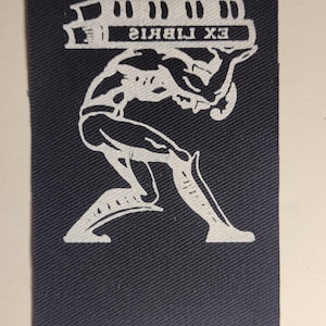 May include: A white patch on a dark blue fabric background with a stylised image of a person carrying a stack of books. The text "EX LIBRIS" is printed on the books.
