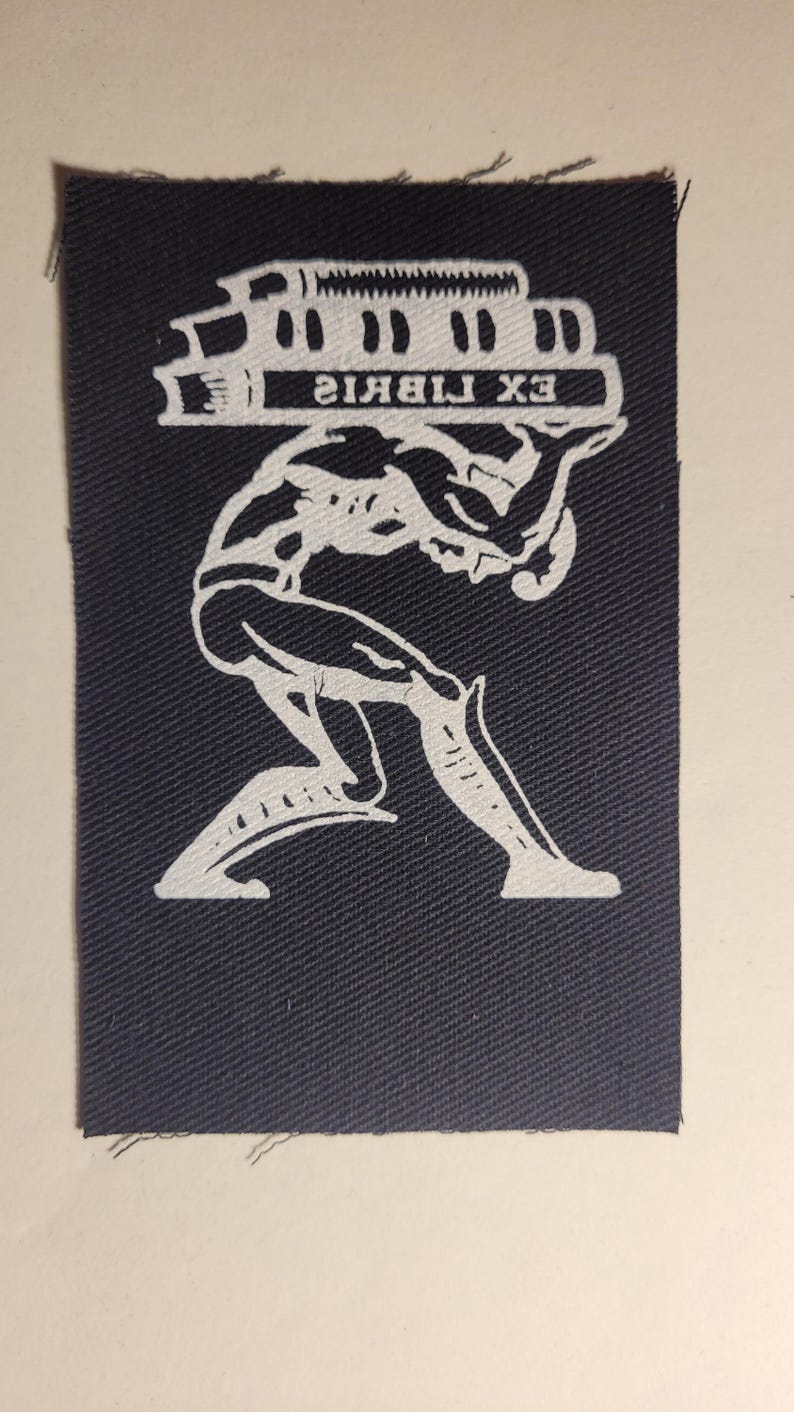 Puede incluir: Un parche blanco sobre un fondo de tela azul oscuro con una imagen estilizada de una persona que lleva una pila de libros. El texto "EX LIBRIS" est&aacute; impreso en los libros.