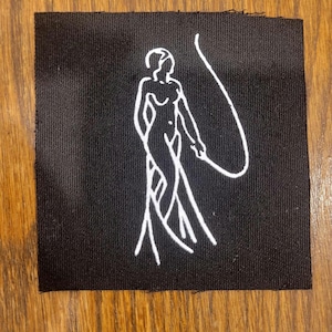 May include: A square black fabric patch featuring a white line art illustration of a standing figure holding a long, curved whip. The design is centered on the patch, which has a visible woven texture and straight, slightly frayed edges.