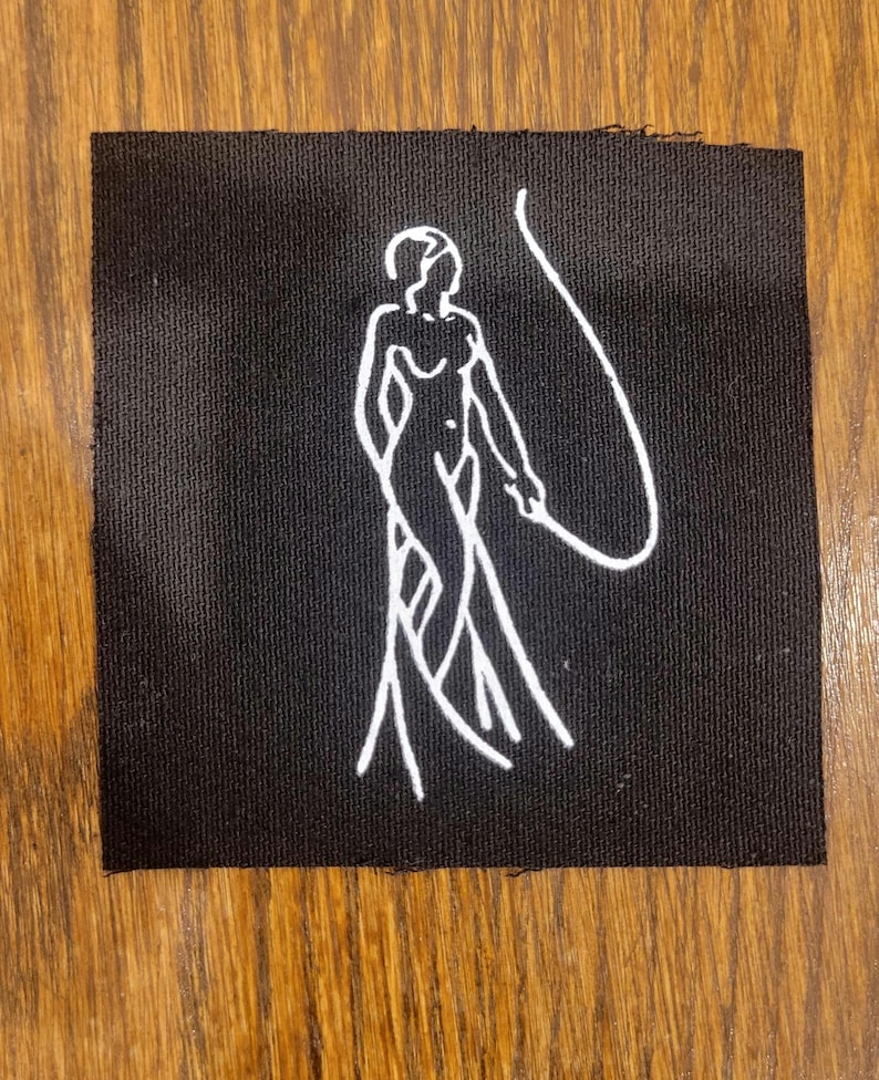 Puede incluir: Un parche de tela negro cuadrado que presenta una ilustraci&oacute;n de l&iacute;neas blancas de una figura de pie sosteniendo un l&aacute;tigo largo y curvo. El dise&ntilde;o est&aacute; centrado en el parche, que tiene una textura tejida visible y bordes rectos ligeramente deshilachados.