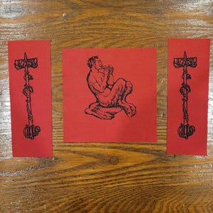 May include: Three red cards with black illustrations. The center card features a satyr playing a flute. The flanking cards display a staff with a decorative top and a spiraling vine design. The cards are arranged on a wooden surface.