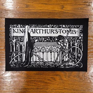 Puede incluir: Parche rectangular negro con ilustración y texto en blanco. El texto dice "KING ARTHUR'S TOMB" sobre una ilustración de una tumba, una figura y árboles. El parche es probablemente para una chaqueta o bolso.