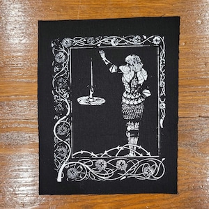 May include: A rectangular black fabric patch featuring a white screen-printed illustration of a figure in armour reaching towards a sword held by a hand emerging from water, all enclosed within a decorative border of intertwined vines and flowers.