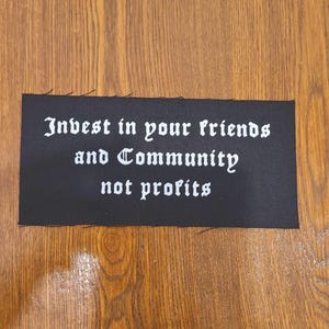 May include: A rectangular black fabric patch with unfinished edges featuring white text in a gothic-style font that reads: Invest in your friends and Community not profits. The patch is a flat, woven textile item with a clear, high-contrast design.