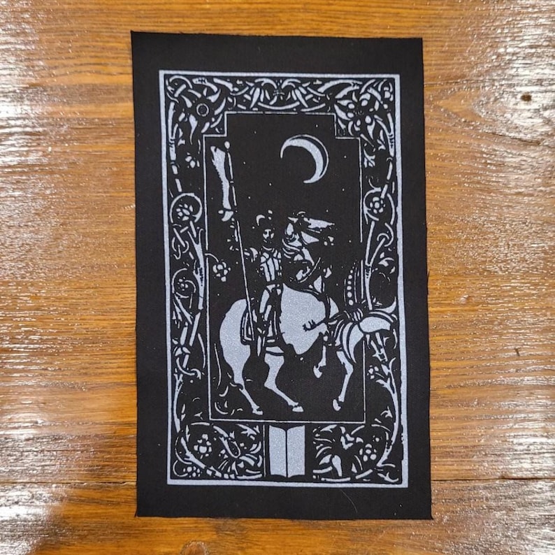 May include: Black and white patch depicting a knight on horseback beneath a crescent moon. The design includes ornate floral and scrollwork borders, and an open book at the bottom. The knight is holding a flag.