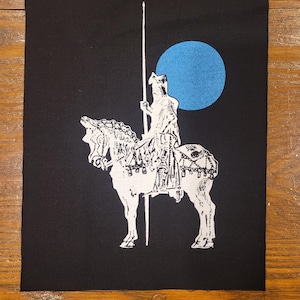 May include: Black fabric patch with a white illustration of a knight on horseback, holding a spear. A blue circle is in the background. The patch is rectangular and made of a woven material.