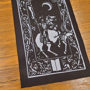 May include: Black rectangular patch with a white illustration of a knight on horseback beneath a crescent moon. The design includes floral and scrollwork borders, and an open book at the bottom. Suitable for clothing or accessories.