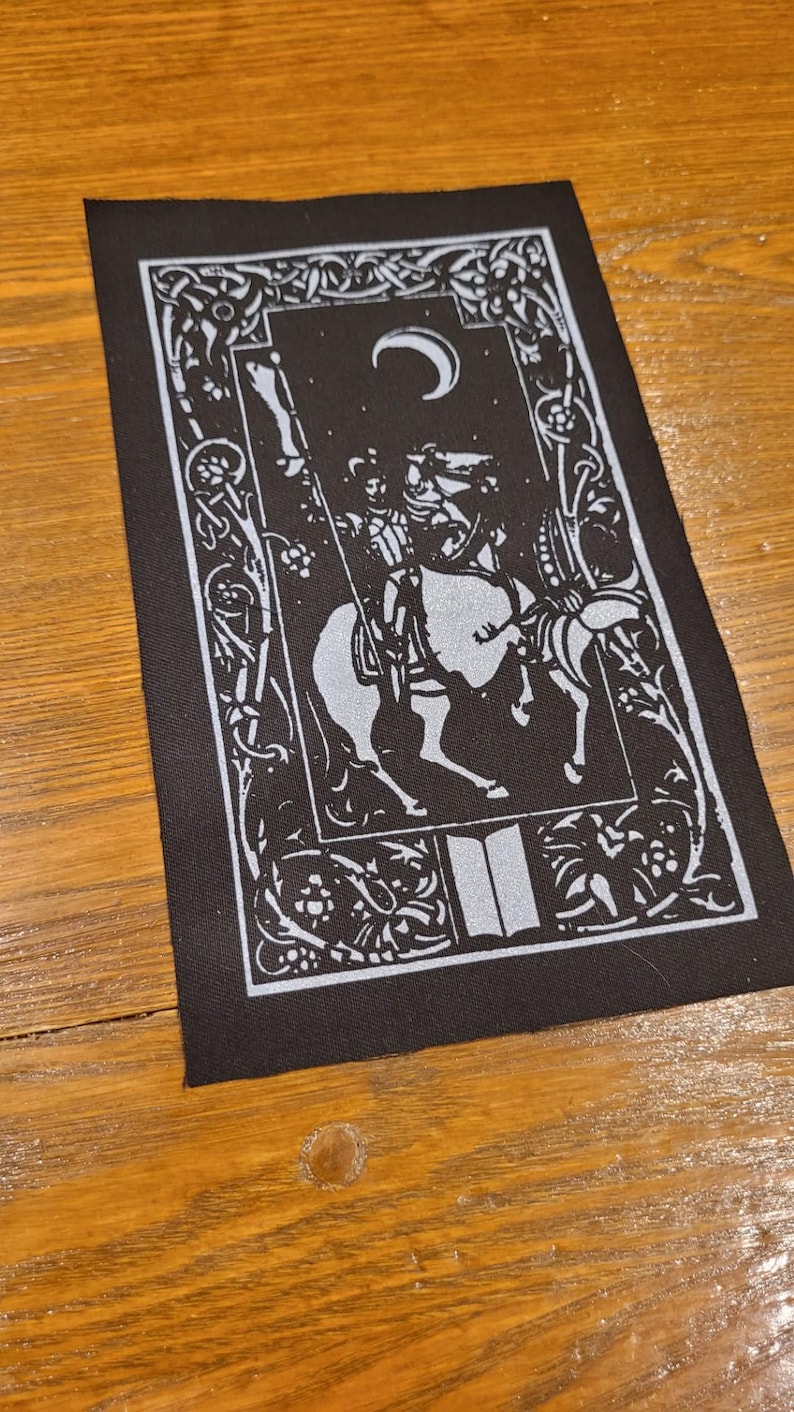 May include: Black rectangular patch with a white illustration of a knight on horseback beneath a crescent moon. The design includes floral and scrollwork borders, and an open book at the bottom. Suitable for clothing or accessories.