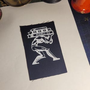 Puede incluir: Un parche de tela negro con una imagen blanca de una persona que lleva una pila de libros. El texto "EX LIBRIS" est&aacute; impreso en los libros.