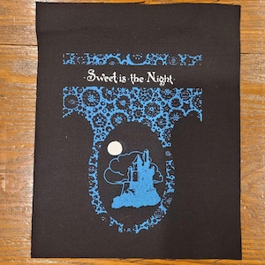 May include: Black fabric patch with the text "Sweet is the Night" in white script. The patch features a blue design of a castle, moon, and clouds. The design is framed by a blue floral border.