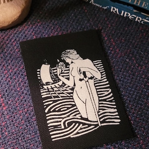 May include: Black and white embroidered patch featuring a woman standing in the water holding a sword. The woman is nude and has long hair. The water is depicted with wavy lines. The patch is rectangular and has a black background.