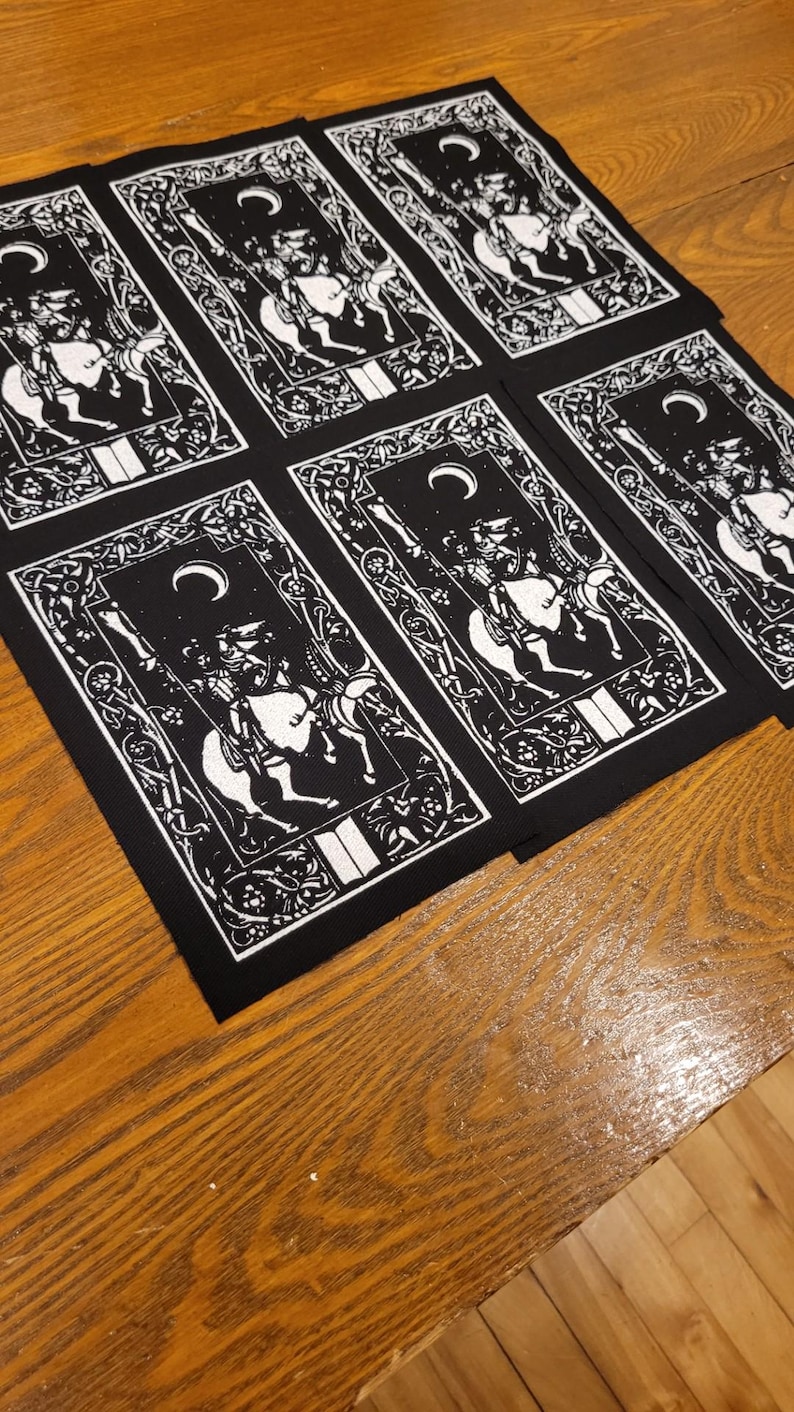 May include: A collection of black and white tarot card patches, each depicting a knight on horseback beneath a crescent moon. The patches have decorative borders and are laid out on a wooden surface. The design is repeated.