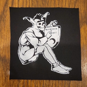 May include: A square black fabric patch featuring a white screen-printed graphic of a seated jester with a skull face. The jester wears a multi-pointed cap with bells and points toward a shield-shaped sign that reads memento mori with a crescent moon.
