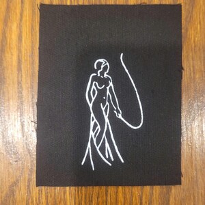 Puede incluir: Un parche rectangular de tela negra con una ilustraci&oacute;n bordada en hilo blanco de una figura de pie que sostiene un l&aacute;tigo largo y curvado. El dise&ntilde;o est&aacute; centrado sobre el tejido oscuro, mostrando una est&eacute;tica minimalista y limpia.