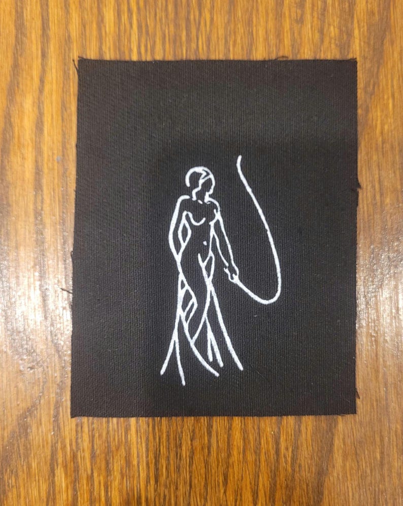 Puede incluir: Un parche rectangular de tela negra con una ilustraci&oacute;n bordada en hilo blanco de una figura de pie que sostiene un l&aacute;tigo largo y curvado. El dise&ntilde;o est&aacute; centrado sobre el tejido oscuro, mostrando una est&eacute;tica minimalista y limpia.