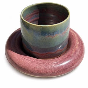 May include: A ceramic cup and saucer set. The cup features a cylindrical shape with a multi-colored glaze in shades of blue, green, and purple. The saucer has a wide, rounded rim with a textured, speckled pink and mauve glaze finish.