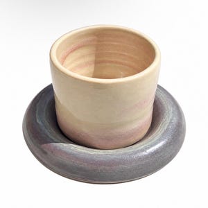 Könnte beinhalten: Eine Keramiktasse mit einer glatten, cremefarbenen und zartrosa glasierten Oberfläche, die mittig auf einer breiten, kreisförmigen Untertasse mit einer gesprenkelten, dunkelgrauen und violetten Glasur und glänzender Textur steht.