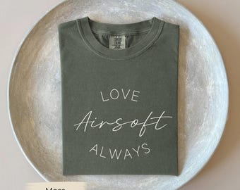 May include: A moss green t-shirt with the words "LOVE Airsoft ALWAYS" in white script. The shirt is folded neatly on a round, silver-colored tray, set against a neutral background. The shirt has a crew neck and short sleeves.