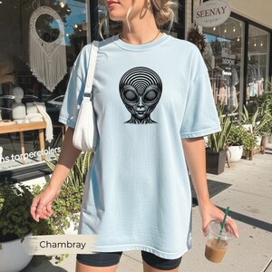 Könnte beinhalten: Hellblaues T-Shirt mit einem schwarz-weißen Alien-Gesichtsmotiv. Der Außerirdische hat große Augen und ein detailliertes Kopfdesign. Das Shirt hat eine lockere Passform und kurze Ärmel. Das Wort "Chambray" befindet sich am unteren Bildrand.
