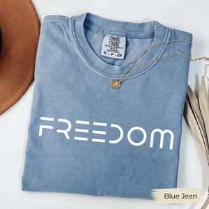 May include: A blue jean Comfort Colors t-shirt with the word "FREEDOM" printed in white. A gold necklace with a pendant is draped across the shirt. The shirt is folded on a white surface.