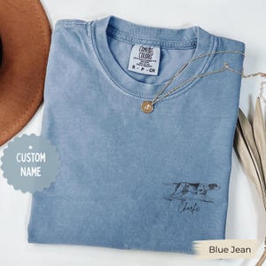 Puede incluir: Camiseta Comfort Colors azul vaquero con un gráfico de perro y el nombre "Charlie" impreso debajo. Un collar dorado con un colgante descansa sobre la camiseta. Talla pequeña.