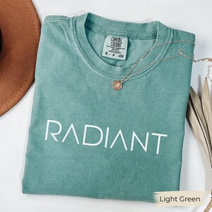 A light green t-shirt with the word "RADIANT" printed on it, along with a necklace and a brown hat, all laid out on a white surface.