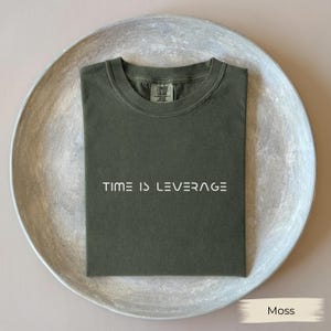 May include: A moss green t-shirt with the text "TIME IS LEVERAGE" in white. The shirt is folded neatly on a silver-colored, round tray. The shirt is made of a soft, comfortable material.