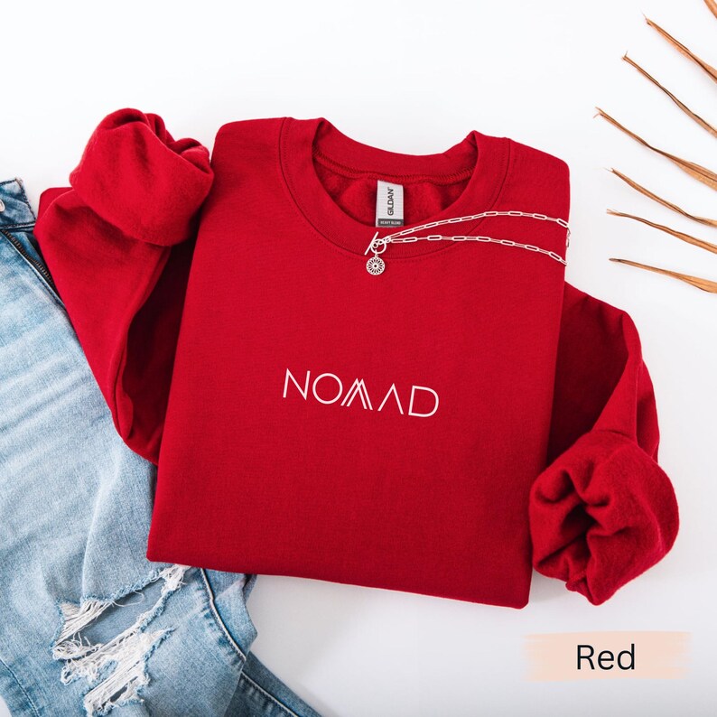 A red sweater with the word "NOMAD" printed on it, along with a necklace and some denim jeans, are laid out on a white background.