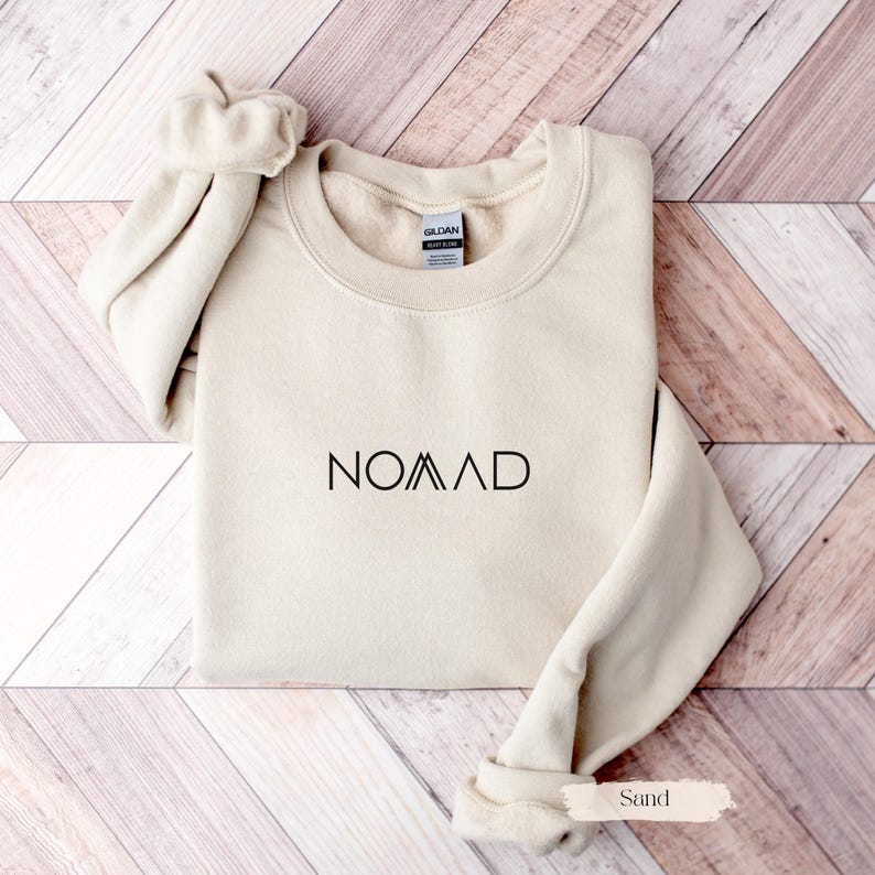 A beige sweater with the word "NOMAD" printed on it, laid out on a wooden surface.