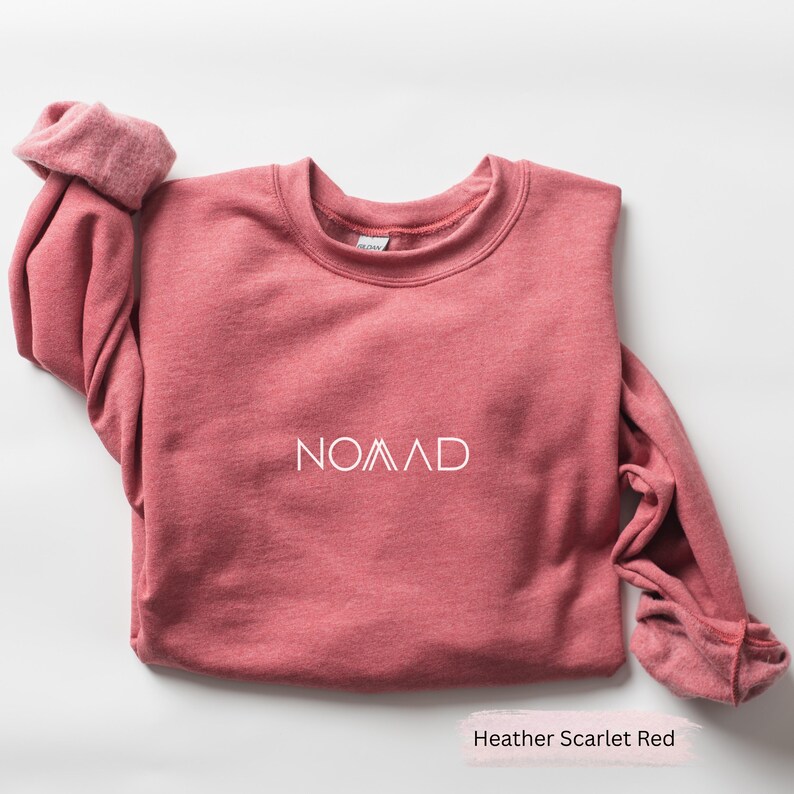 A red sweater with the word "NOMAD" printed on it, along with a tag, is folded and placed on a white surface.