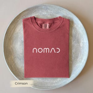 A red t-shirt with the word "NOMAD" printed on it, laid out on a gray plate.