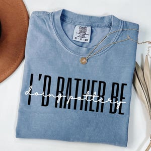 Puede incluir: Una camiseta azul Comfort Colors con el texto "I'd rather be doing pottery" en blanco y negro. Un collar dorado con un colgante está sobre la camiseta.