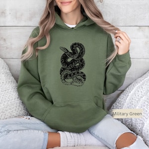 Puede incluir: Sudadera con capucha verde militar con un diseño de serpiente negra. El diseño de la serpiente es detallado y estilizado. La sudadera tiene una capucha con cordón y un bolsillo de canguro. El color se describe como "Verde militar".