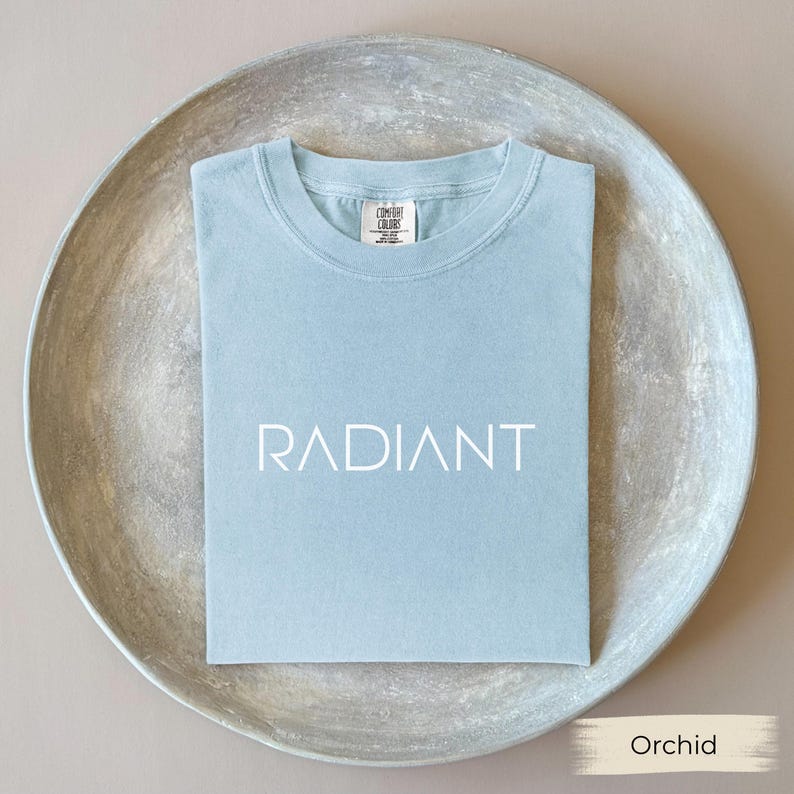 A light blue t-shirt with the word "RADIANT" printed on it, laid out on a round, textured plate.