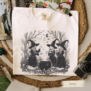 May include: Ivory-colored t-shirt featuring a black and white illustration of three witches gathered around a cauldron in a forest setting. The witches wear pointed hats and dresses. The shirt has a Comfort Colors tag.