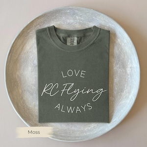 May include: A moss green t-shirt with the words "LOVE RC Flying ALWAYS" in white script. The shirt is folded neatly on a round, light gray tray. The shirt is made of a soft, comfortable material.