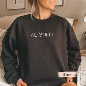 May include: Black sweatshirt with the word "ALIGNED" in white, centered on the chest. The sweatshirt has a crew neck and long sleeves. The word "Black" is in a white box at the bottom right of the image.